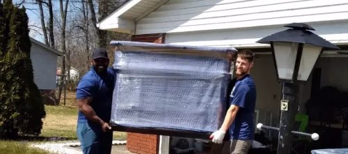 Two men moving wrapped furniture outdoors.
